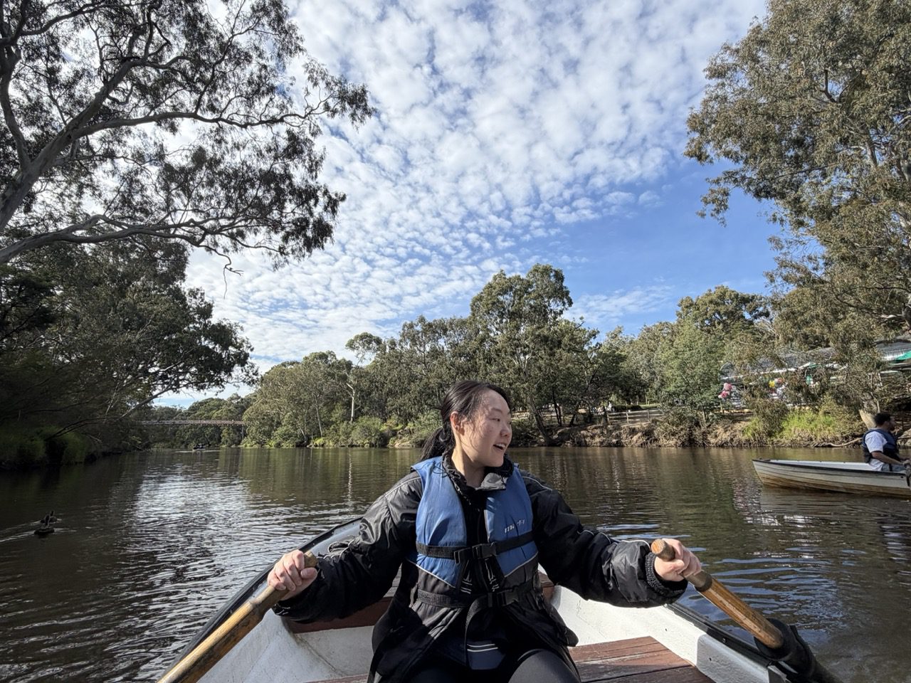 A woman sits in a rowing boat, holding two wooden oars and wearing a blue life vest. A blue sky and trees surround the image.